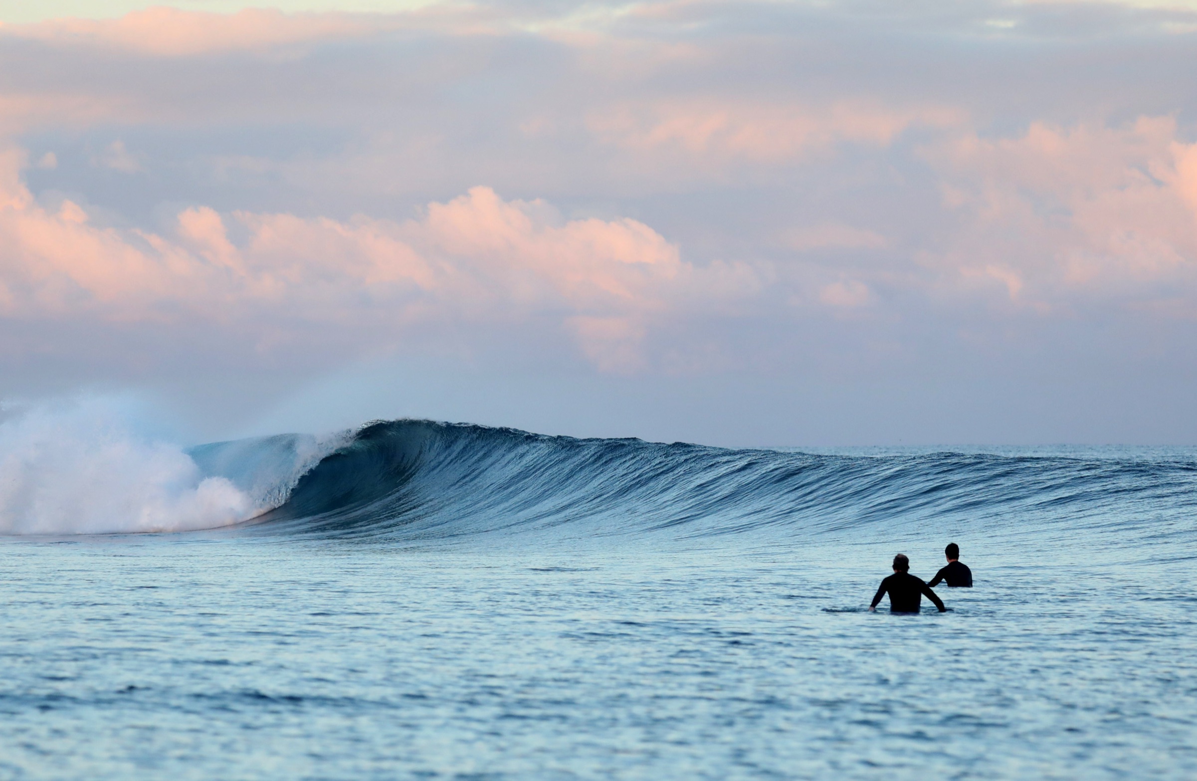 Two surfers waiting in the lineup at dawn as a clean wave peels behind them